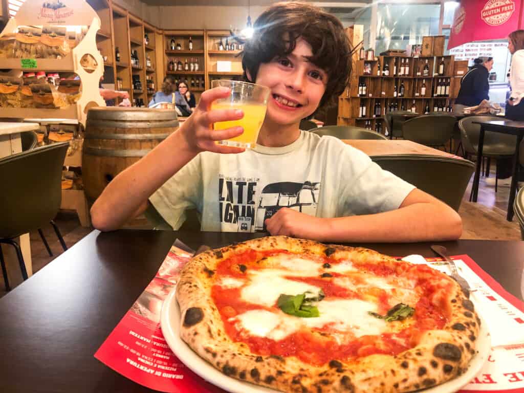 side view of boy holding a drink with a large pizza on the table in front of him.