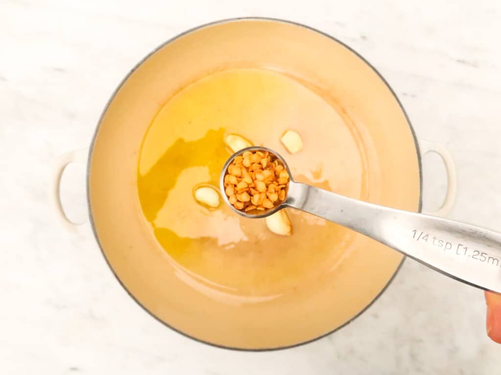 Metal measuring spoon with small sand colored spice over pot with oil and garlic cloves.