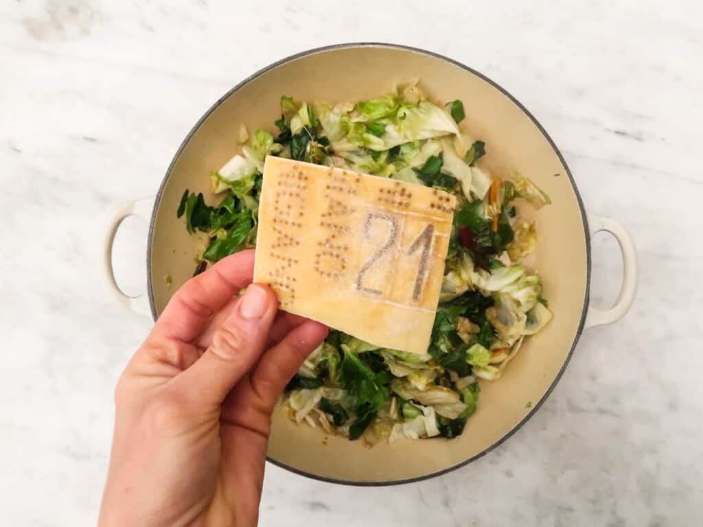 Hand holds parmesan rind over a pot of greens.