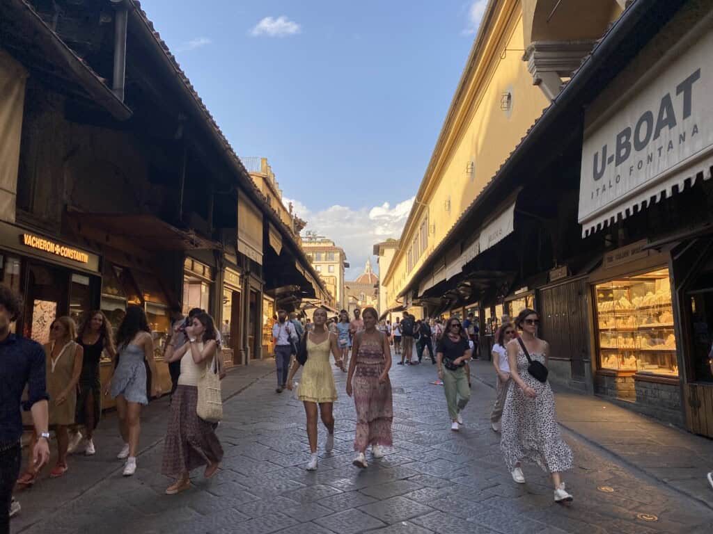 People walk across the Ponte Vecchio in Florence, Italy on a sunny day. Gold shops are open and their products are displayed behind glass on either side of the bridge.