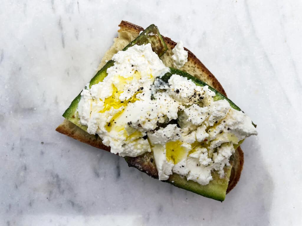 Toasted bread with zucchini and soft italian cheese with pepper and drizzled olive oil, sitting on a white marble surface.