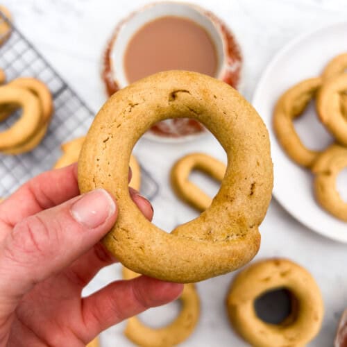Hand holds up an 'o' shaped butter cookie above a white counter and other cookies cooling and a tea cup of hot cocoa.