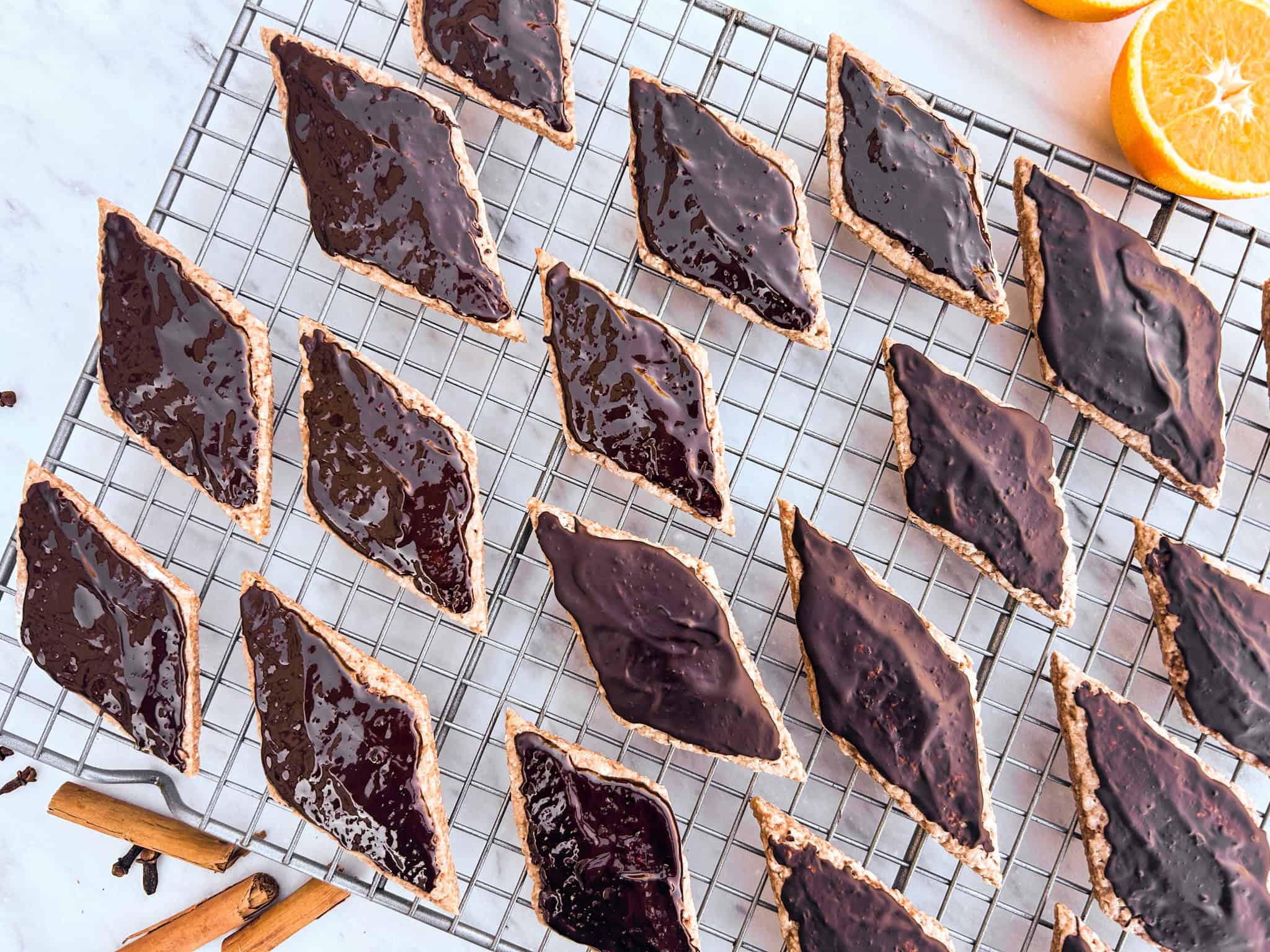 Mostaccioli cookies with chocolate on top. They're sitting on top of a metal cooling rack. Sliced oranges and cinnamon sticks next to rack.