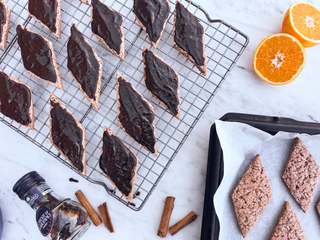Cooling rack with mostaccioli cookies and a baking tray next to it with cooked but unfrosted mostaccioli. Halved orange in upper right corner and spices in lower left.