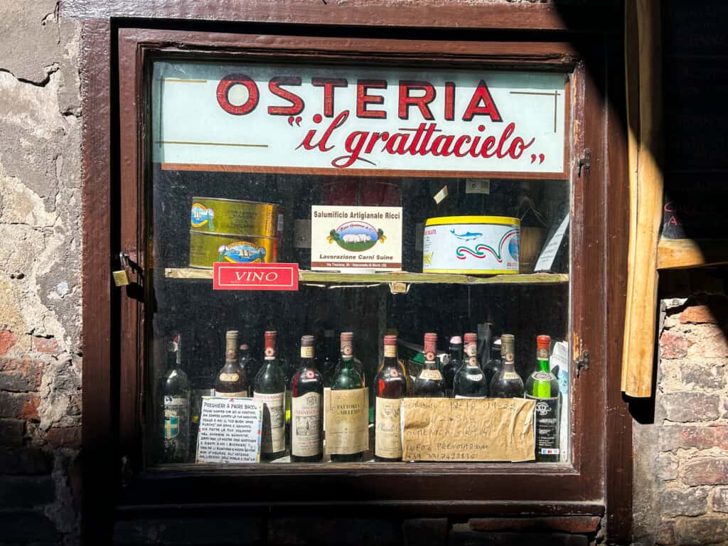 Window of the Osteria il Grattacielo. The name is written in red on a white sign. In the window are large tins of tuna and old bottles of wine.
