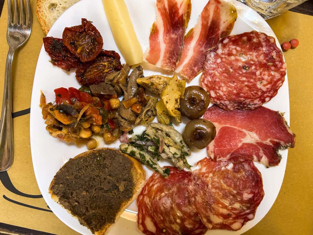 White plate with assorted cured meats, a crostino with liver pate, and vegetables under oil.