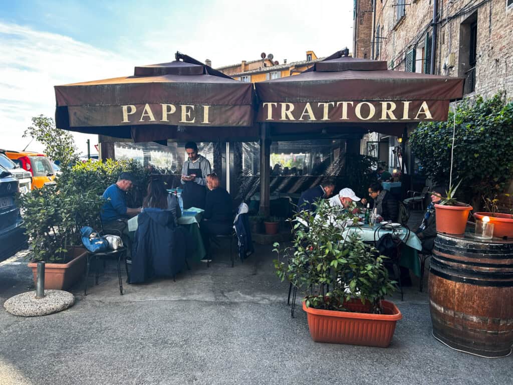 Brown umbrellas hang over tables at an outdoor restaurant in Siena, Italy. The name of the restaurant, Papei Trattoria, is stamped in cream color on the umbrellas. A waiter takes an order from table on left and others eat at tables on right. Plants in planter boxes around the seating area.