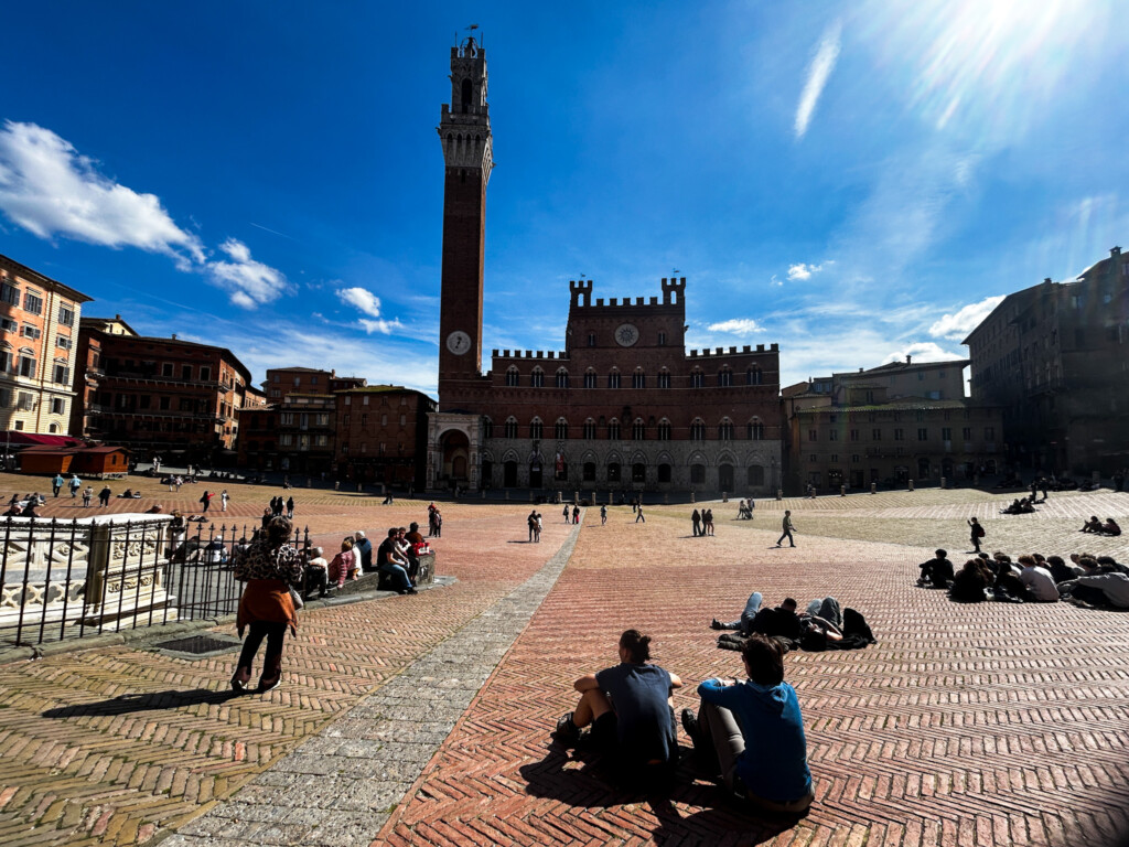 Sunny day in the main piazza of Siena, Italy. People sit in the piazza and look at the clock tower and town hall.