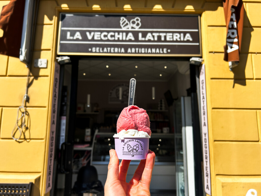 a picture of a hand holding a cup of red and white gelato in front of a yellow building.